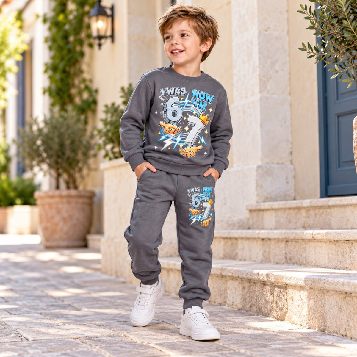 Boy modelling grey I Was 6 Now I'm 7 birthday sweatshirt and matching joggers, full-length standing view
