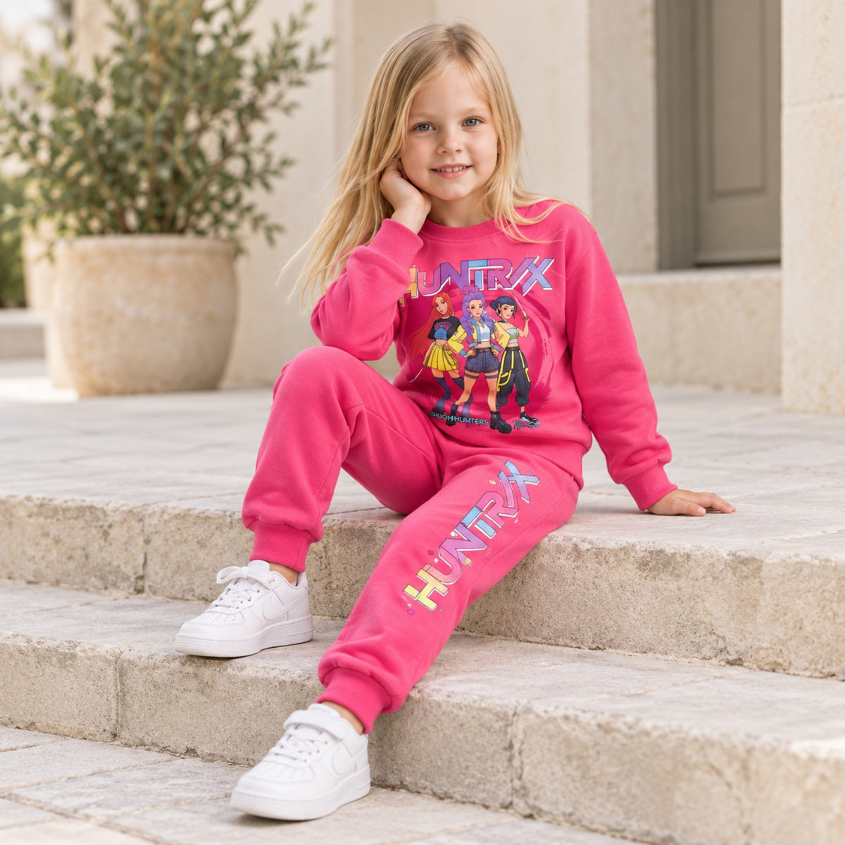 Girl wearing bright pink HUNTR/X matching sweatshirt and joggers sitting on outdoor stone steps